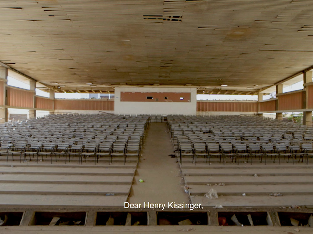 An empty dilapidated hall in Angola filled with chairs. The subtitle reads: 'Dear Henry Kissinger'