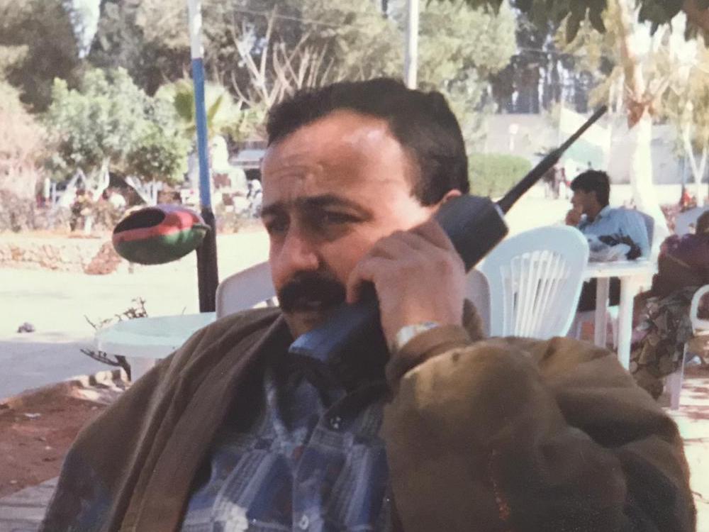 A man on a 90s black brick phone sits on a plastic table outside