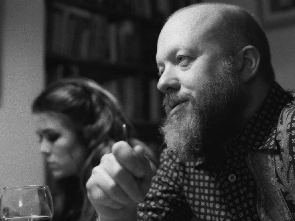 A black and white image of a bald, male presenting person with a beard. He looks off screen to the lefthand is holding a fork