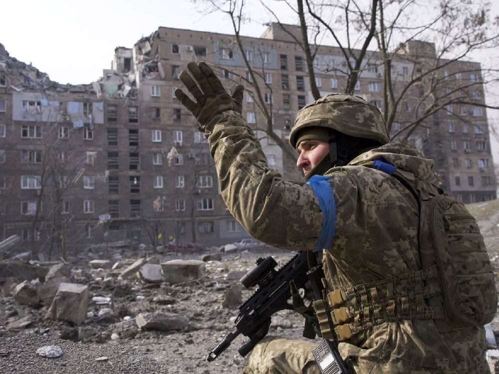 a soldier kneels in front of a half destroyed housing building, holding a machine gun.