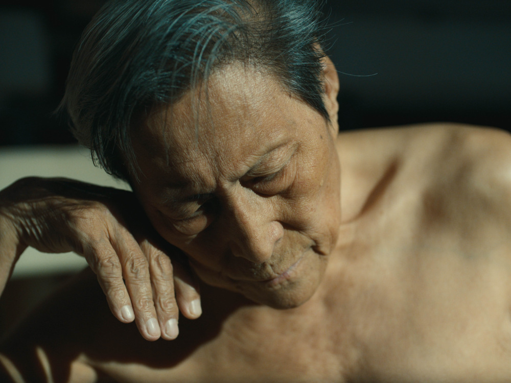 An elderly Chinese man with no top on rests with his hand up against his cheek.