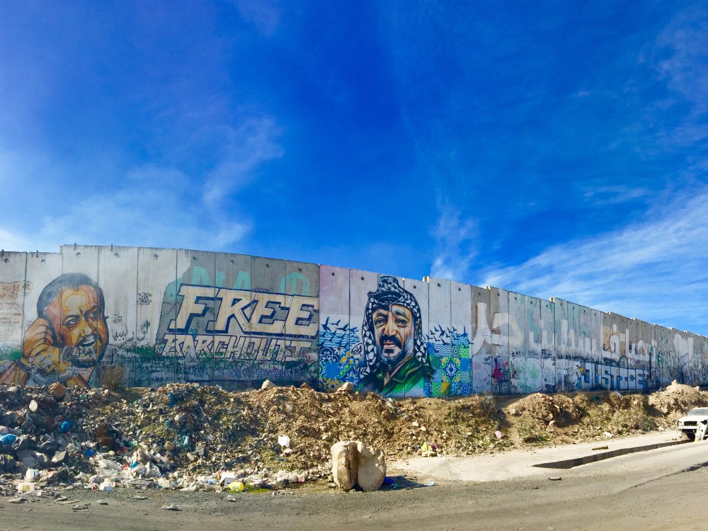 The wall on occupied Palestine, decorated with portraits. Above is a bright blue sky with streaks of clouds stretching through