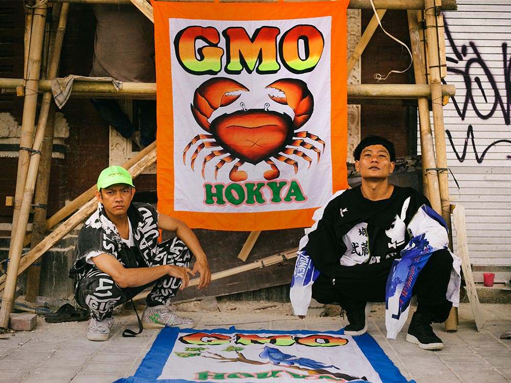 The members of Gabber Modus Operandi, two Indonesian men in graphic printed clothing, sitting in front of fabric hanging from a bamboo structure. The fabric is printed with a crab, and the words 'GMO HOKYA'
