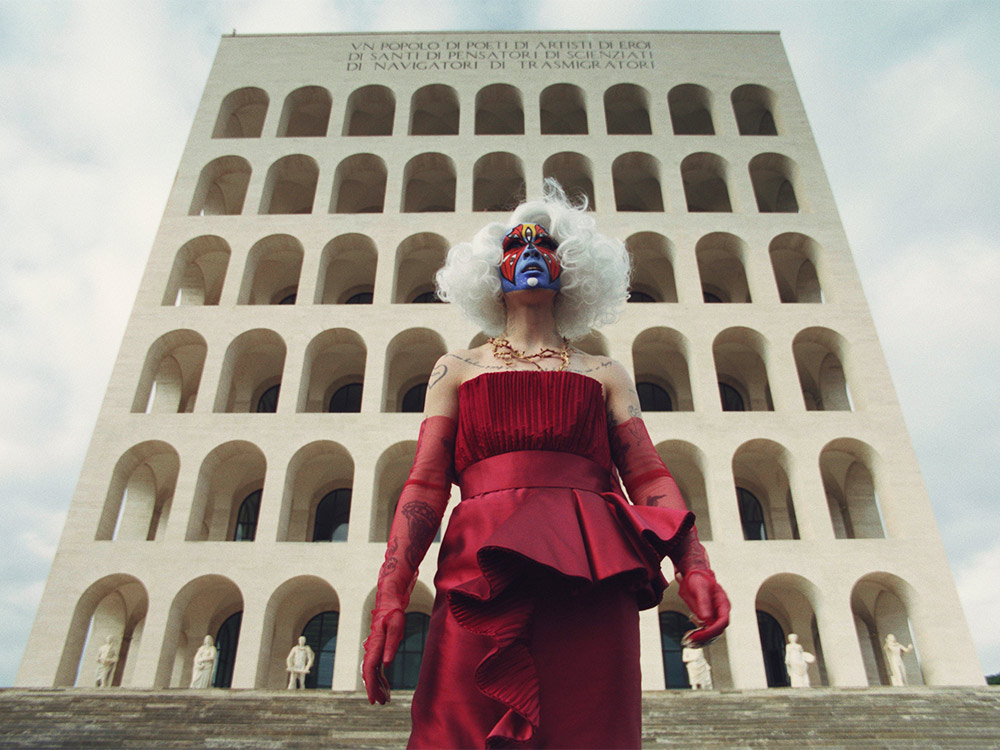 A person in a red dress, platinum blonde hair and blue and red makeup satands in front of a concrete building covered by arches, statues and Latin text