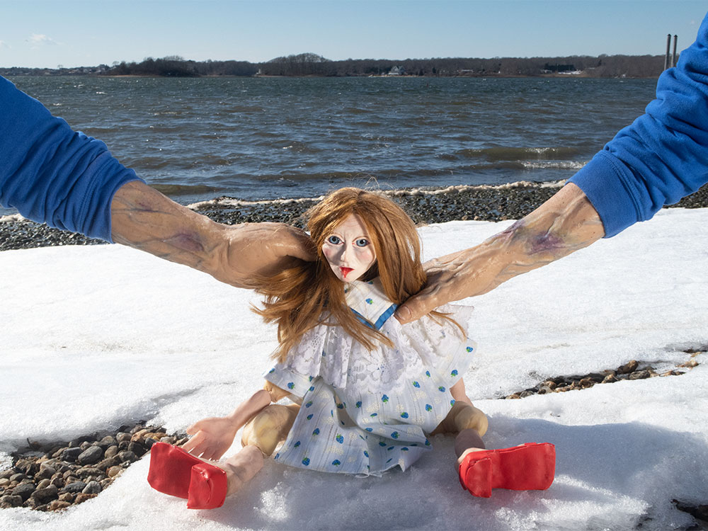 A spooky doll sits on a snow by the water, held by two hands