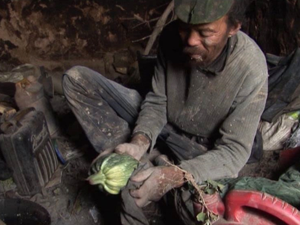 A Chinese man sits in his homemade home, tending to a vegetable. His clothes are covered in dirt