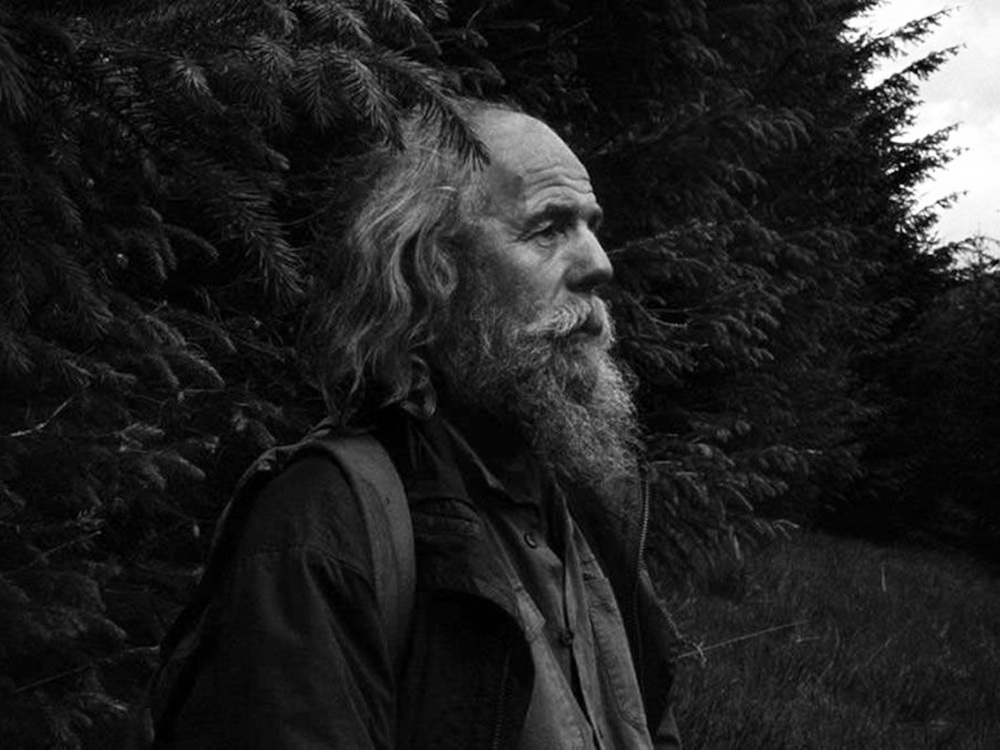 In black and white, a man with long hair and a beard stares off, in a grassy field