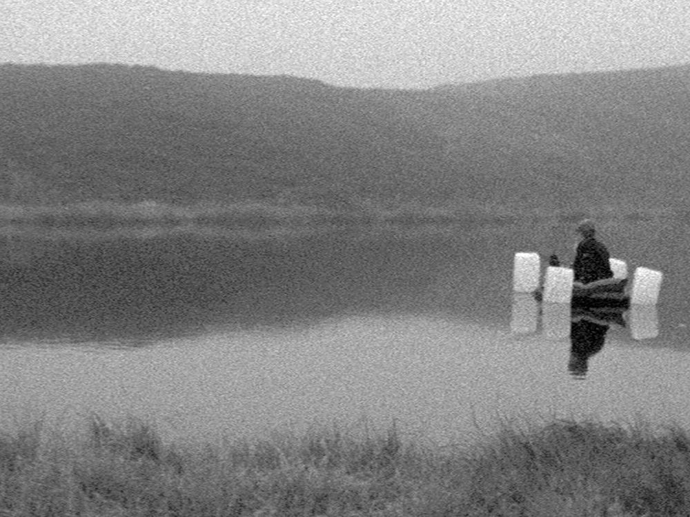 In black and white film, a person sits on a small homemade floatation platform in a river