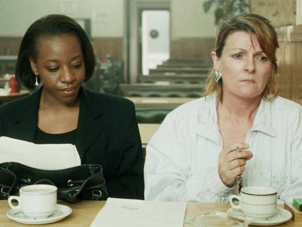 A black woman and a white woman share a coffee. The black woman looks at a document on the table. The white woman smokes, looking distraught