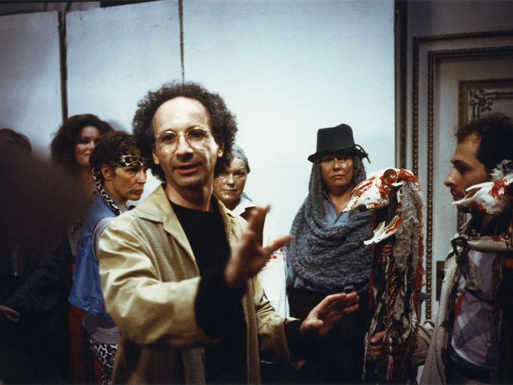 A man with curly hair and glasses stands among a crowd of onlookers, all dressed in unique clothes