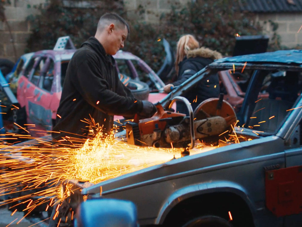 A man in black runs a saw through the front of a car. Sparks are flying