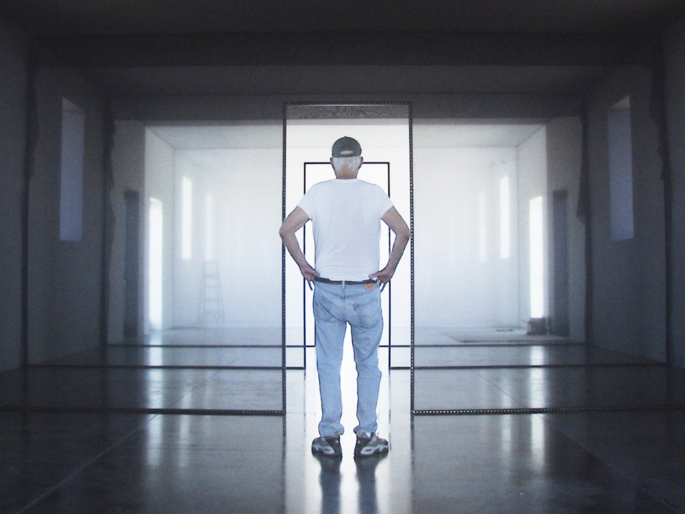 An older man in white shirt and jeans stares down a corridor or frames in a large empty building. Light comes in through the windows
