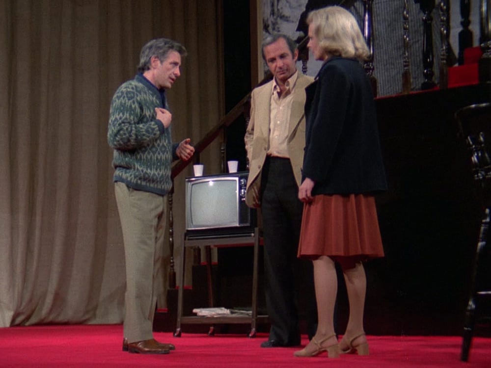 A blonde actress discusses with two formally dressed men, maybe in an argument, in a set with a red velvet floor and a CRT tv