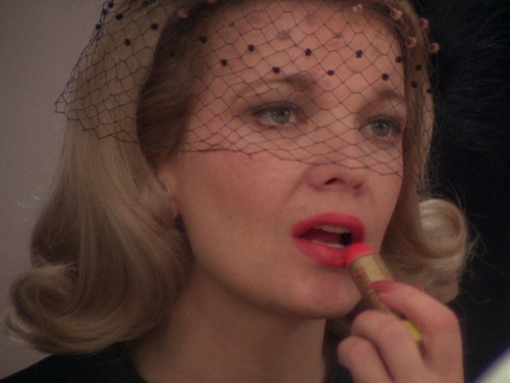 A blonde actress wearing a funeral veil applies bright pink lipstick