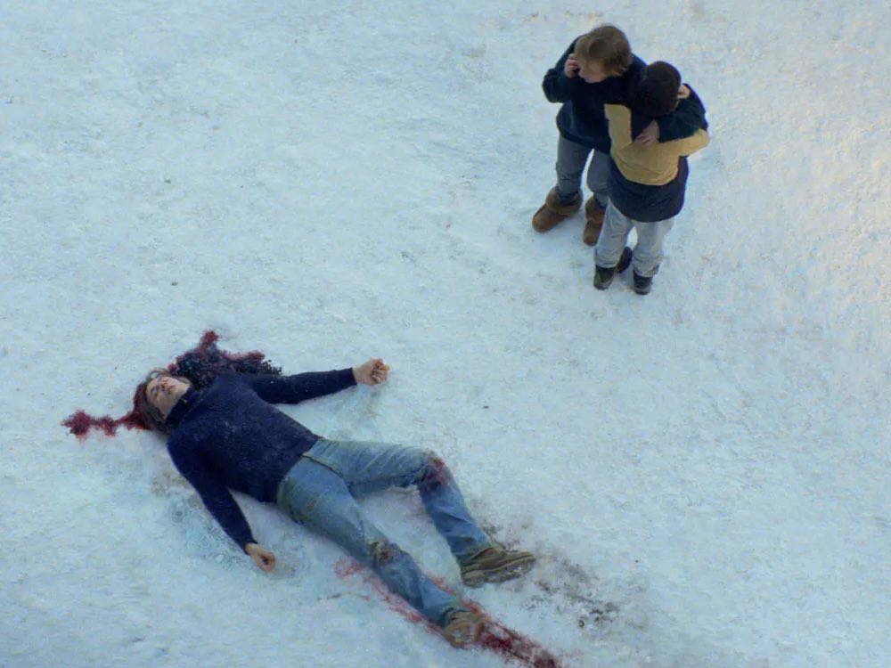 A man rests in the snow with blood coming from his head, and a trail of blood at his feet. His wife and daughter stand by his side, on the phone