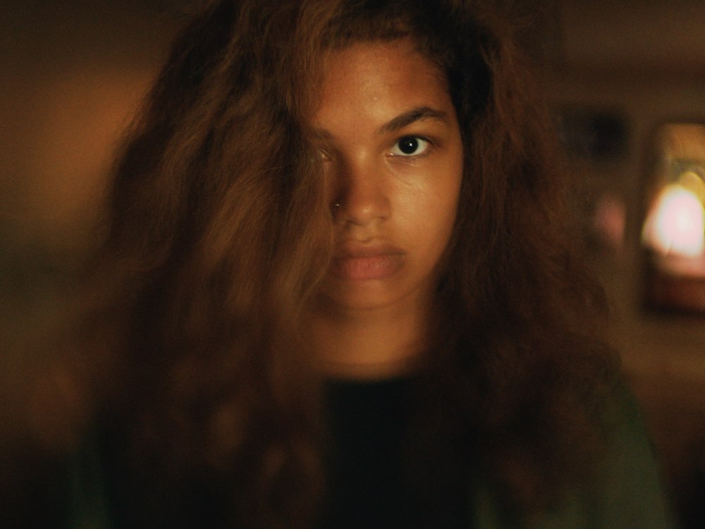 A young black girl looks into the camera, her expression still, hair over half her face. The image is blurred