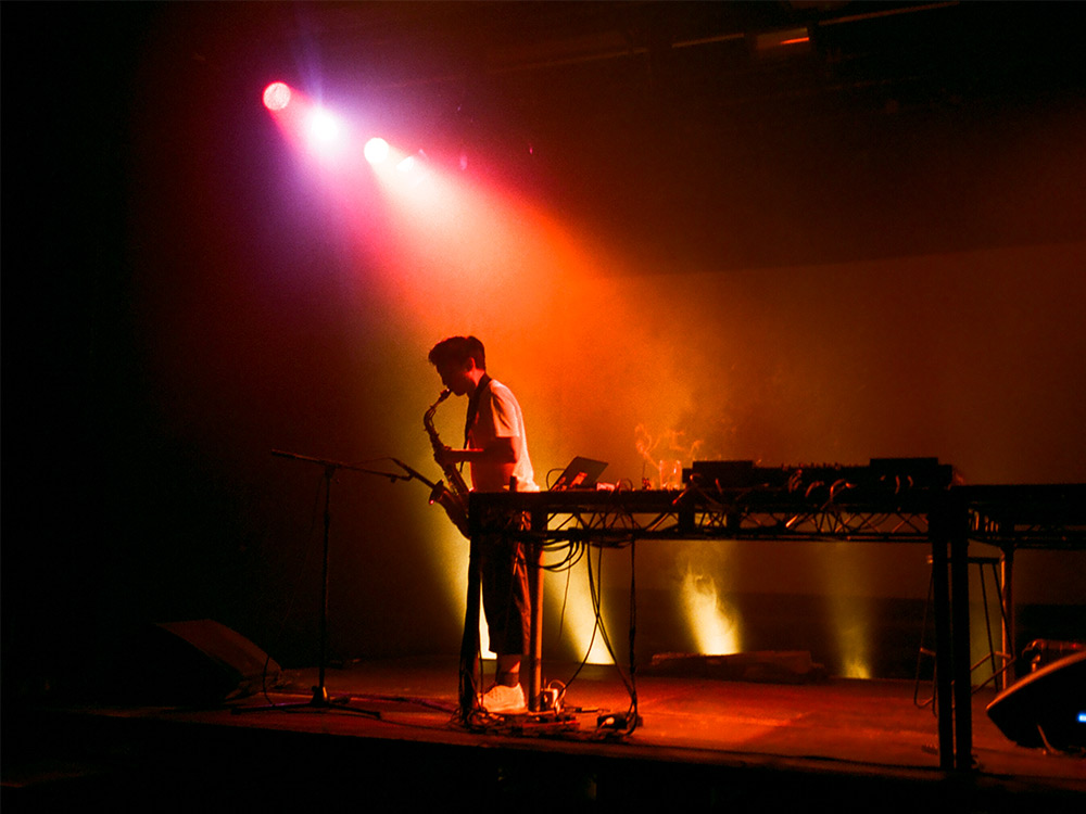 Tzekin playing saxophone on a stage in low orange lights, with incense smoke drifting