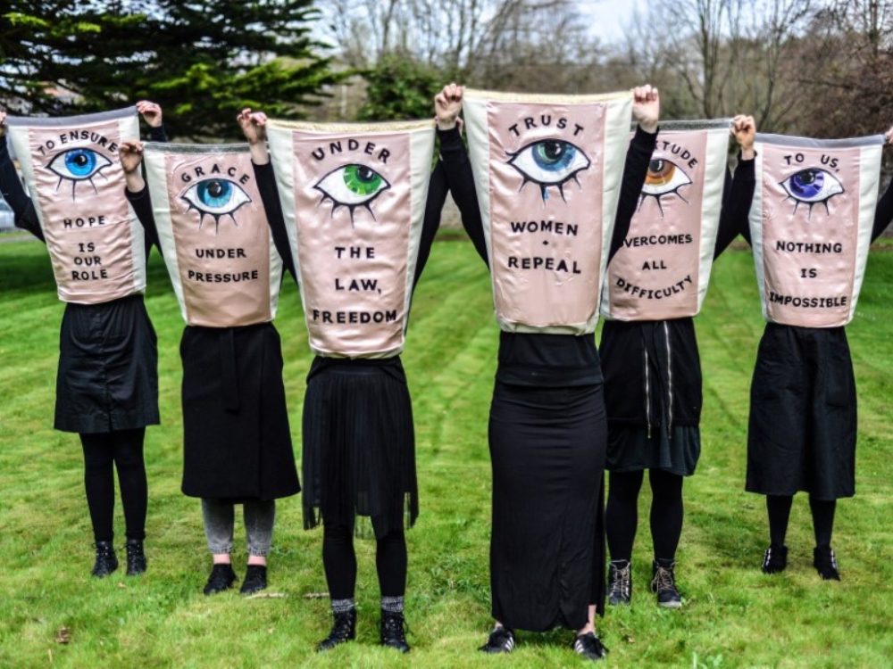 A group of protesters in black carry pink silk banners. They are embroidered with different phrases: 'To ensure hope is our role, Grace under pressure, Trust women repeal, Under the law, freedom - all marked by a different coloured eye