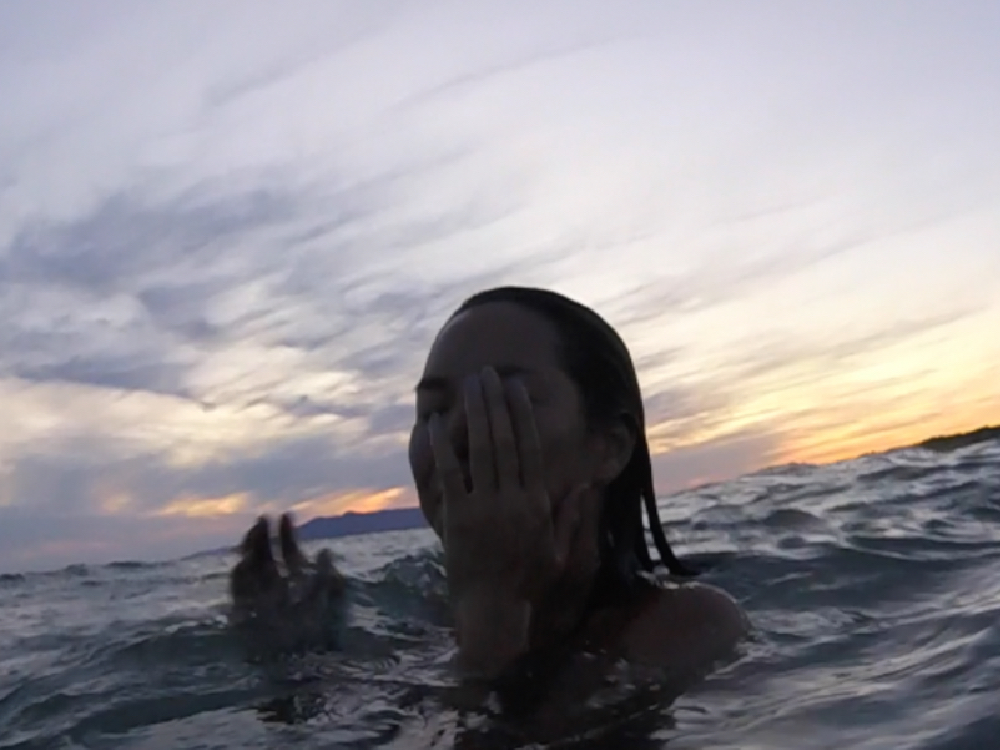 A long haired woman wades through the ocean, her hand over her face, eyes closed, sunset in the background