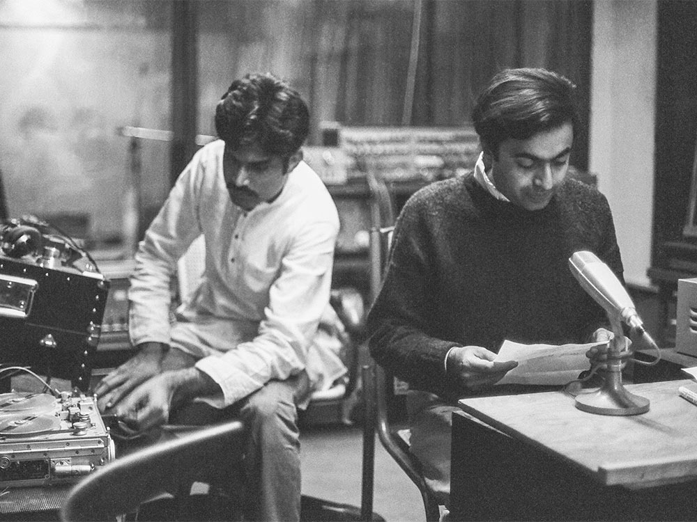 Two south Asian men sit at a recorder and microphone, reading something out on paper