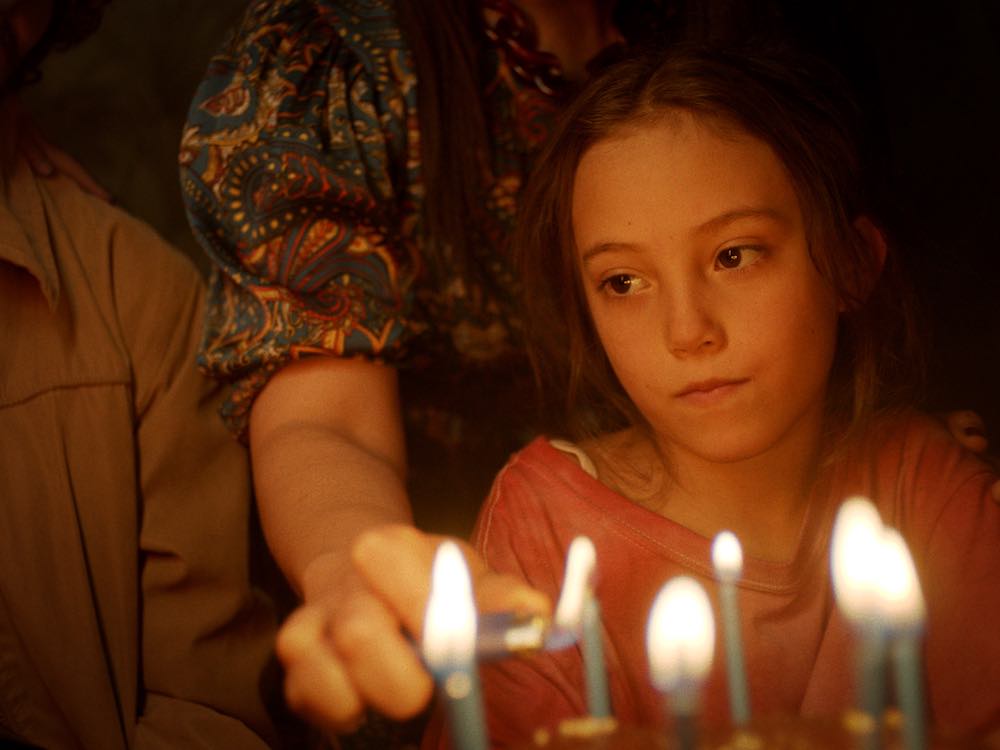 A young girl watches blankly as someone lights candles in front of her