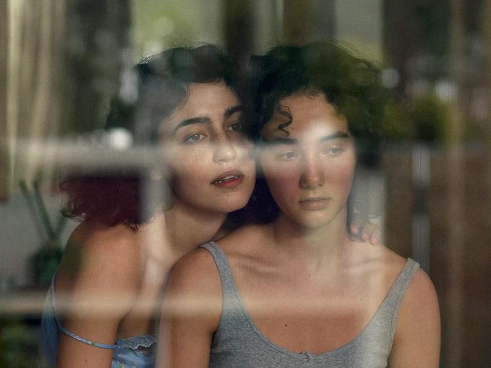 Two women with black curly hair wearing singlets sit looking out the window. One leans over the other with a longing look, while the other looks down, embarassed