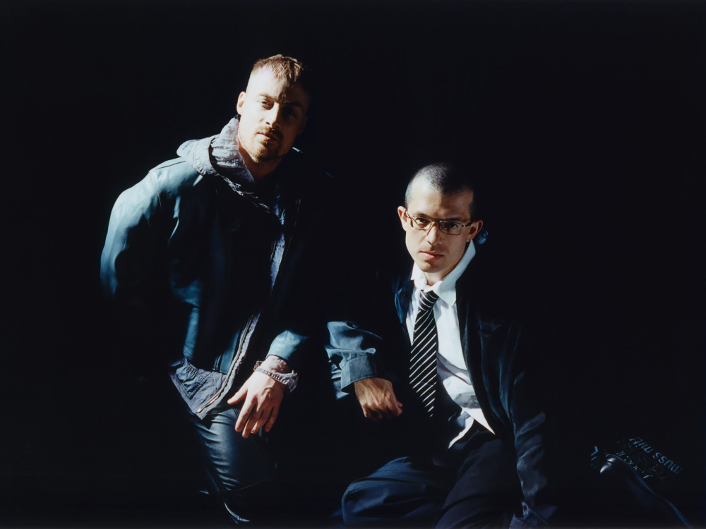 Two white men in formal clothes sit against a black background with bright white light