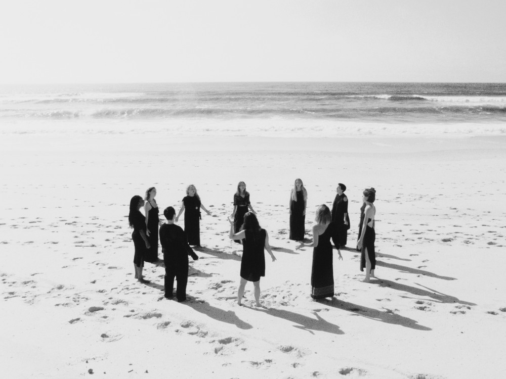 A group wearing black stand in a circle singing on the beach. The photo is in black and white